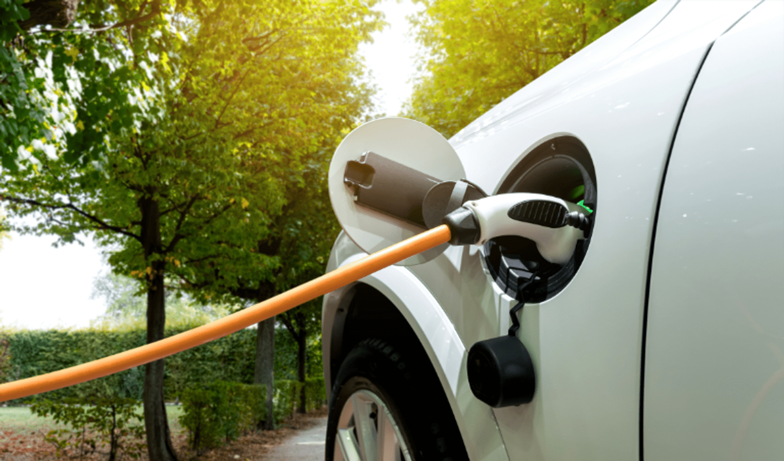 Close-up of an electric vehicle charging at a home EV charging station, with a charging cable connected to the vehicle in a leafy outdoor setting, highlighting clean energy transport and electric vehicle adoption.