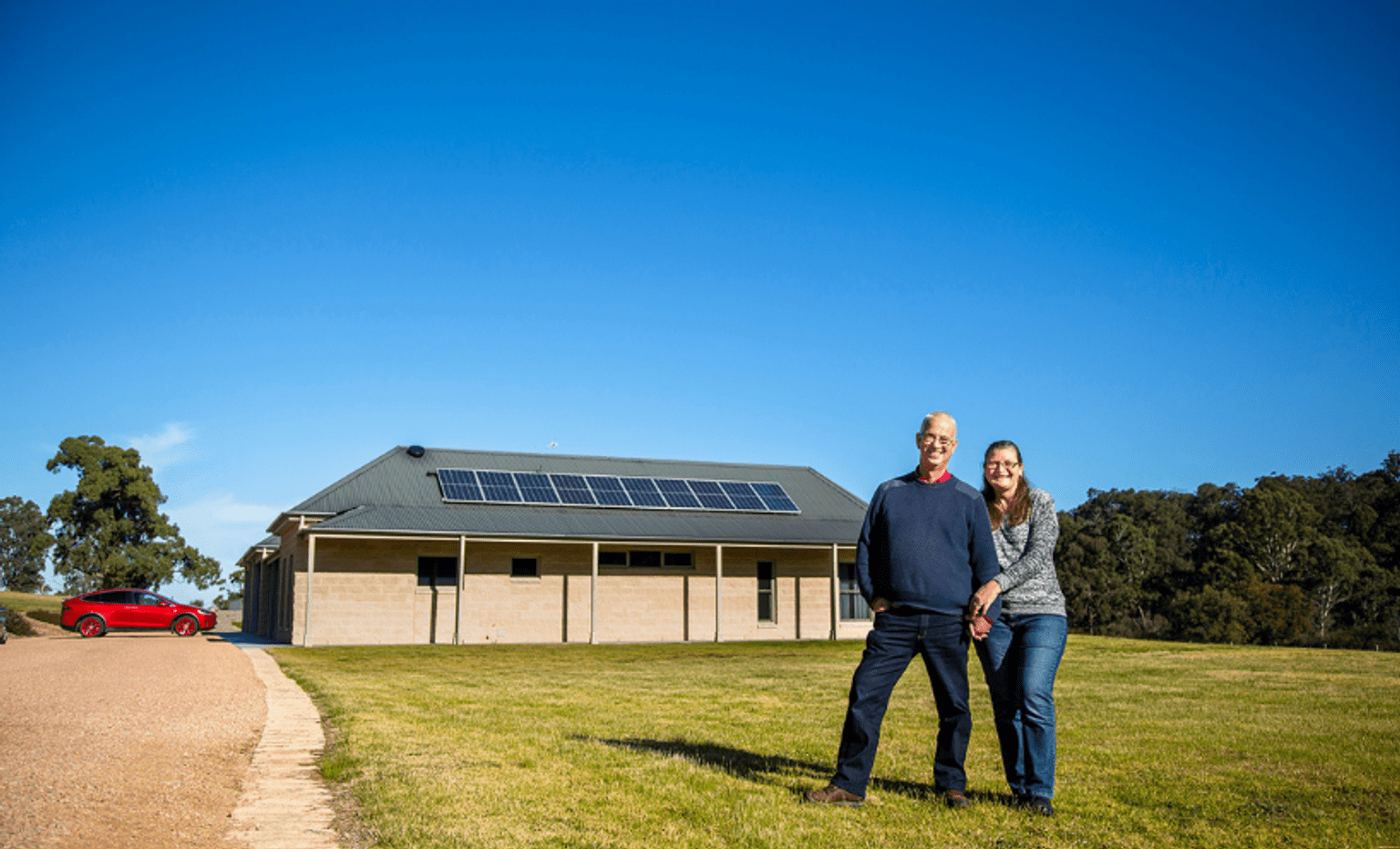 A couple smile at the samera, with their house with solar panels and a red car in the background