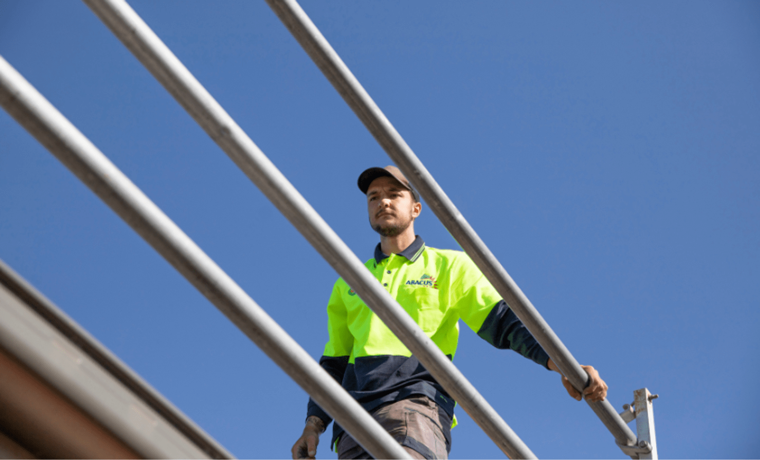 Crackdown on falls risks in the solar sector. A worker in high-visibility safety clothing stands on an elevated structure behind metal guardrails, viewed from below against a clear blue sky, highlighting the importance of fall prevention and safe working practices when working at height.